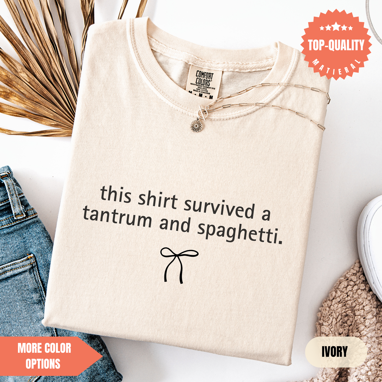 a beige t-shirt with a humorous text printed on it, along with a small bow symbol. The t-shirt is displayed on a white background, and there are some jeans and a sweater visible in the background.