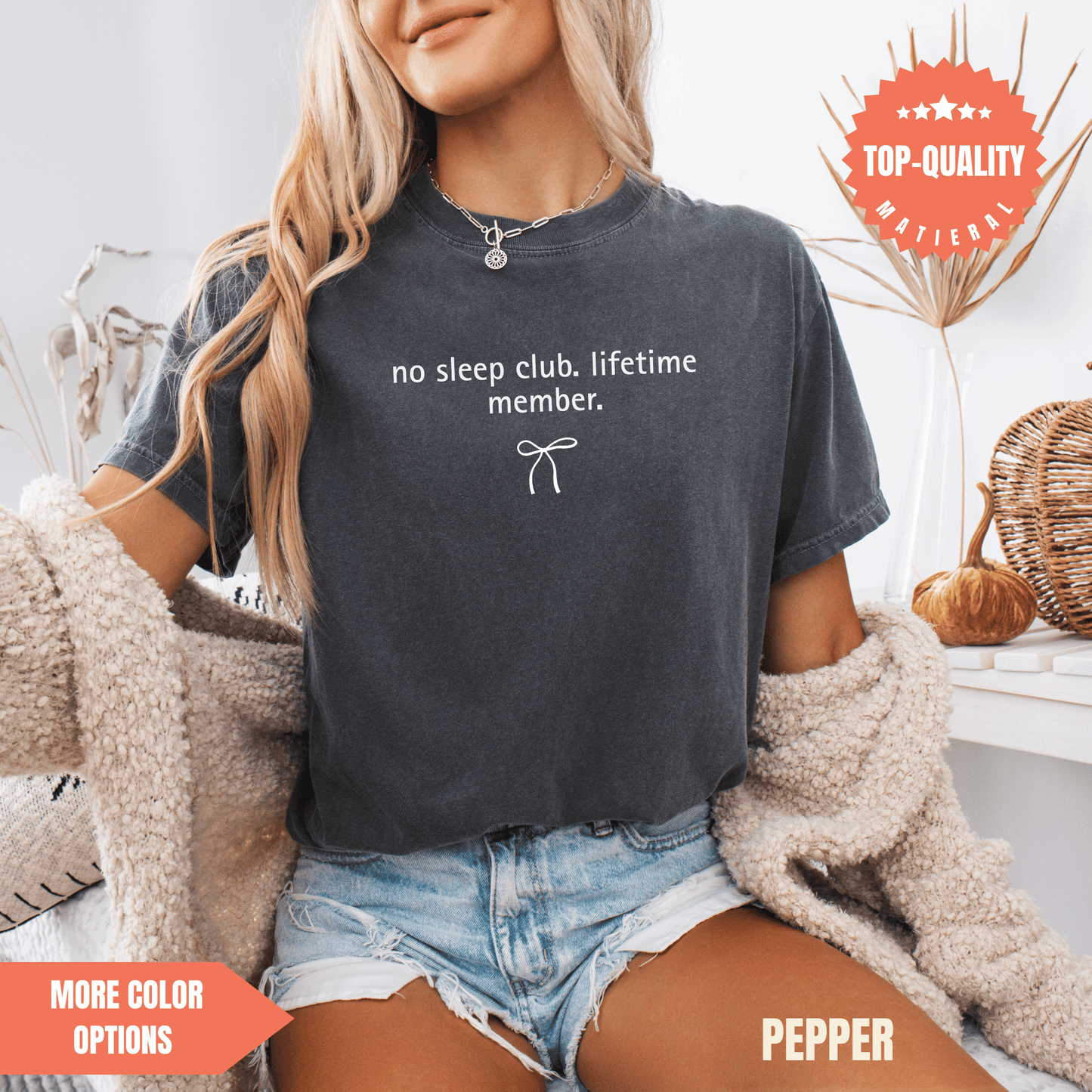 A woman wearing a gray t-shirt with the text "no sleep club lifetime member" printed on it, paired with denim shorts and a beige cardigan.