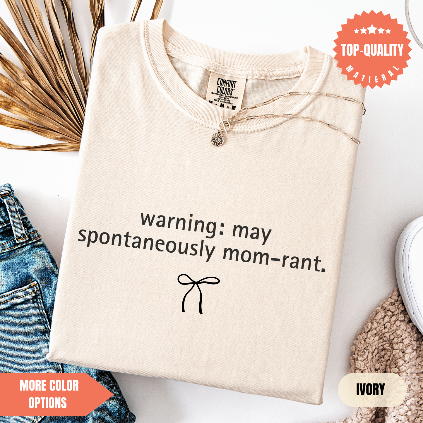 a beige t-shirt with a humorous text printed on it, along with a small bow symbol. The t-shirt is displayed on a white background, and there are some jeans and a sweater visible in the background.