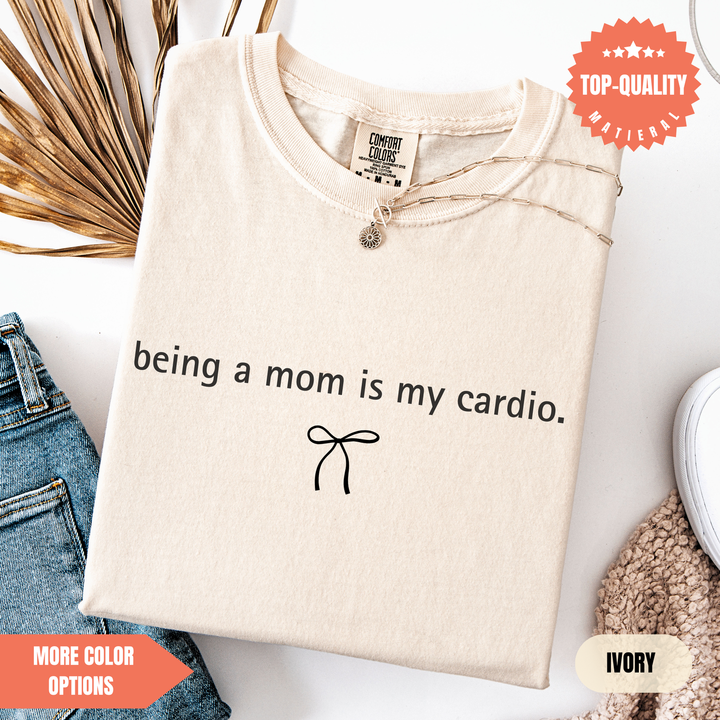 a beige t-shirt with the text "being a mom is my cardio." printed on it, along with a small bow symbol. The t-shirt is displayed on a white background, and there are some jeans and a sweater visible in the background.