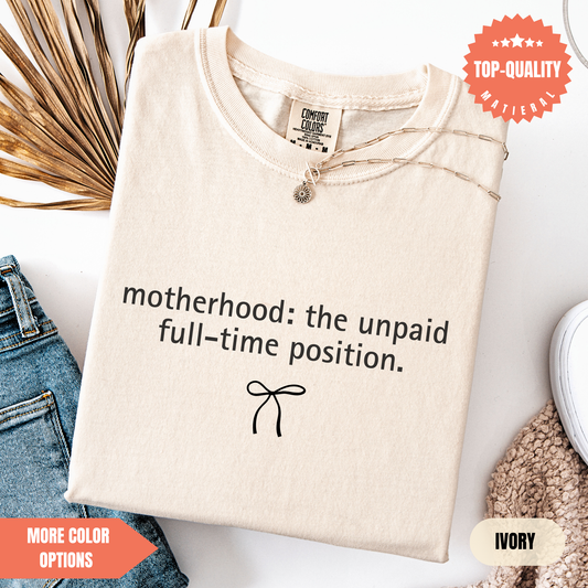 a beige t-shirt with a humorous text printed on it, along with a small bow symbol. The t-shirt is displayed on a white background, and there are some jeans and a sweater visible in the background.