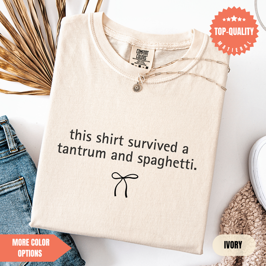 a beige t-shirt with a humorous text printed on it, along with a small bow symbol. The t-shirt is displayed on a white background, and there are some jeans and a sweater visible in the background.