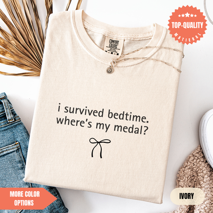 a beige t-shirt with a humorous text printed on it, along with a small bow symbol. The t-shirt is displayed on a white background, and there are some jeans and a sweater visible in the background.