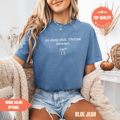 A woman wearing a blue t-shirt with the text "no sleep club, lifetime member." and denim shorts, sitting in a chair with a beige sweater.