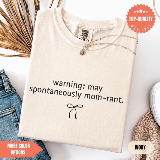 a beige t-shirt with a humorous text printed on it, along with a small bow symbol. The t-shirt is displayed on a white background, and there are some jeans and a sweater visible in the background.