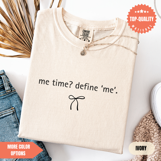 a beige t-shirt with the text "me time? define me'?" printed on it, along with a small bow symbol. The t-shirt is displayed on a white background, and there are some jeans and a sweater visible in the background.