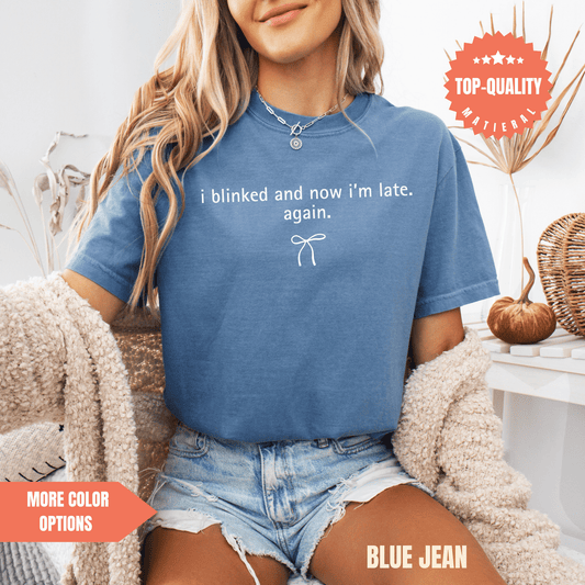 A woman wearing a blue t-shirt with a humorous text and denim shorts is sitting in a chair.